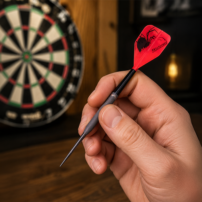 Hand holding a Laserdarts Black Widows Movable Point Smooth Steel Tip with a red and black spider design in front of a dartboard.