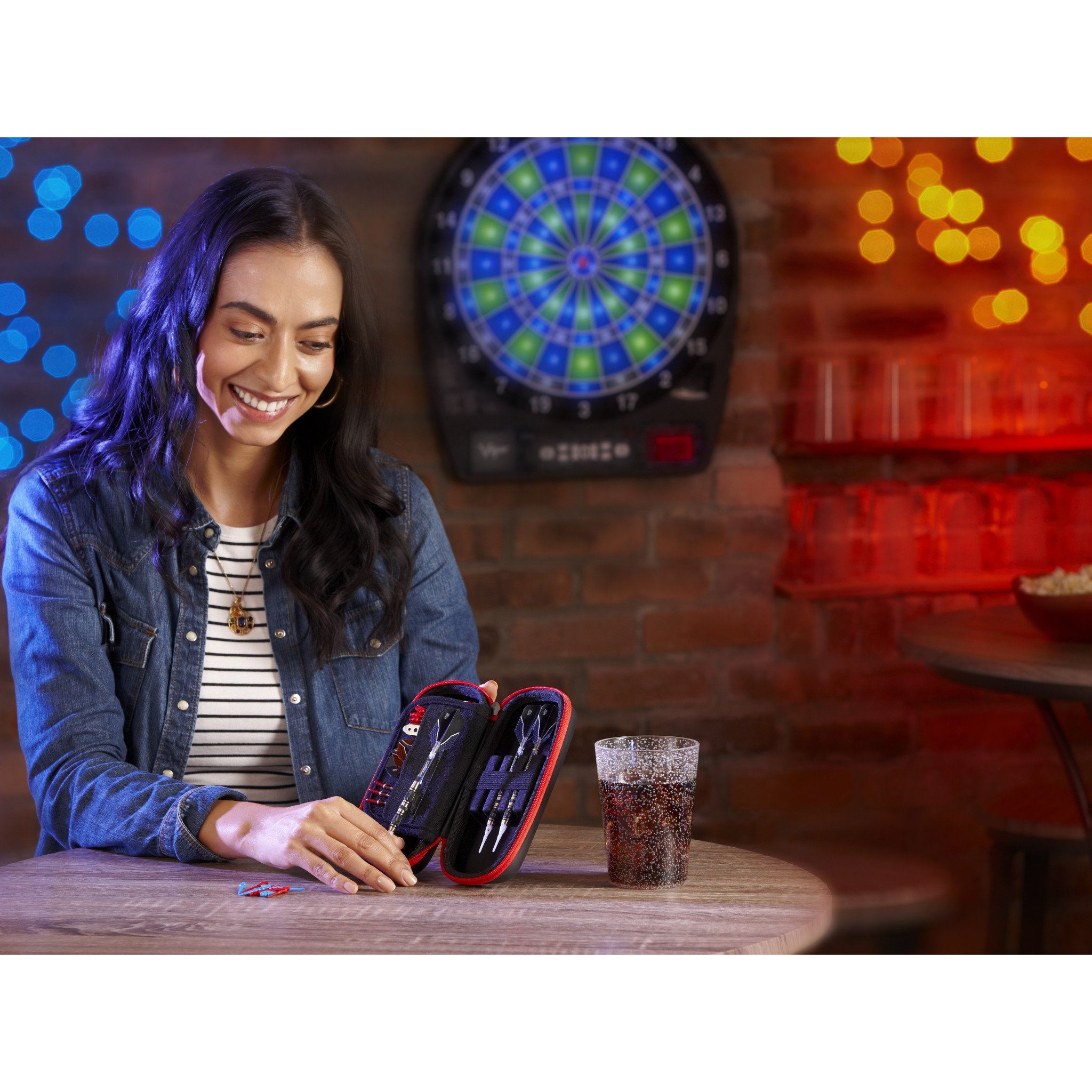 Person opening a red and black dart case at a table with dartboard behind