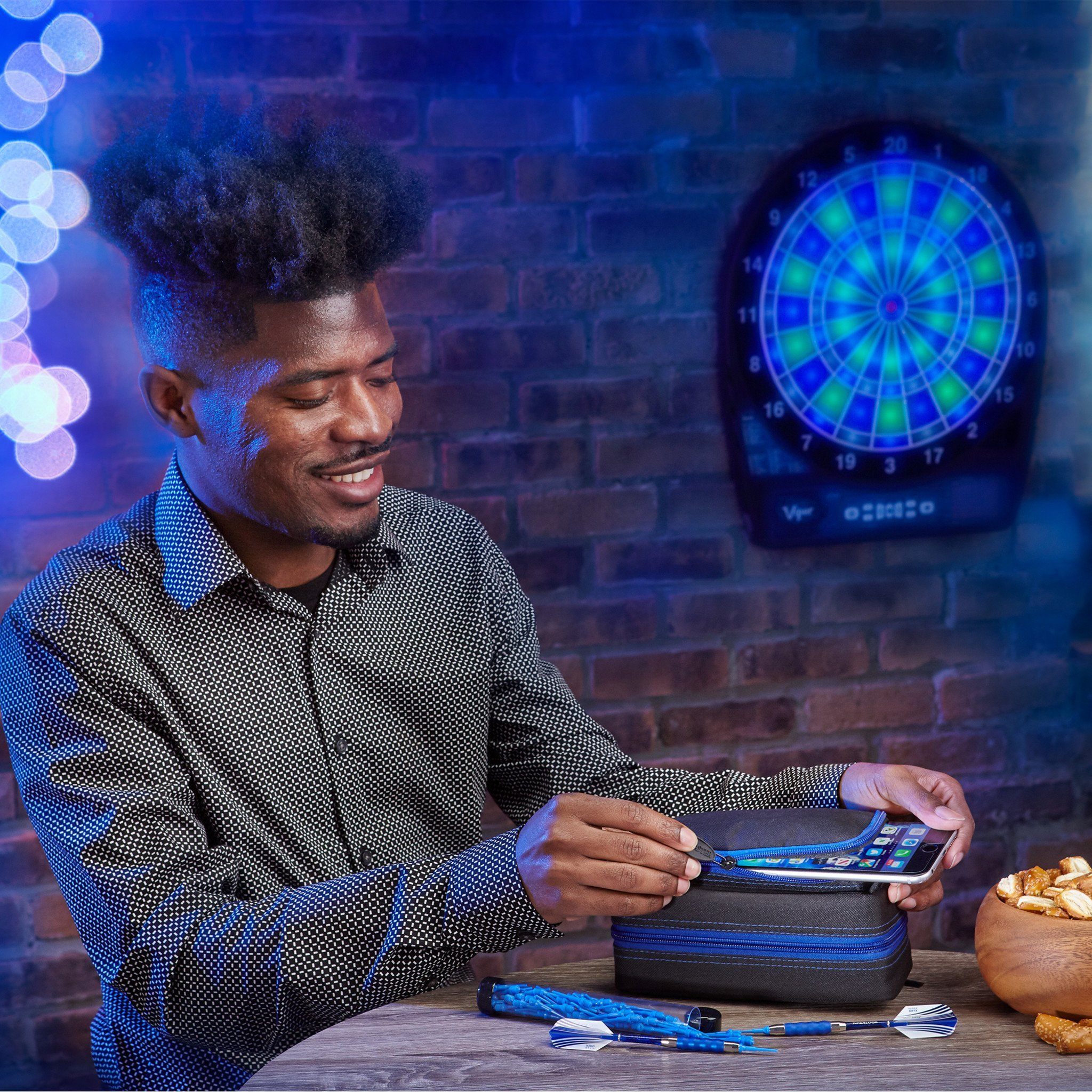 Person holding smartphone next to black dart case with blue zipper and electronic dartboard on wall