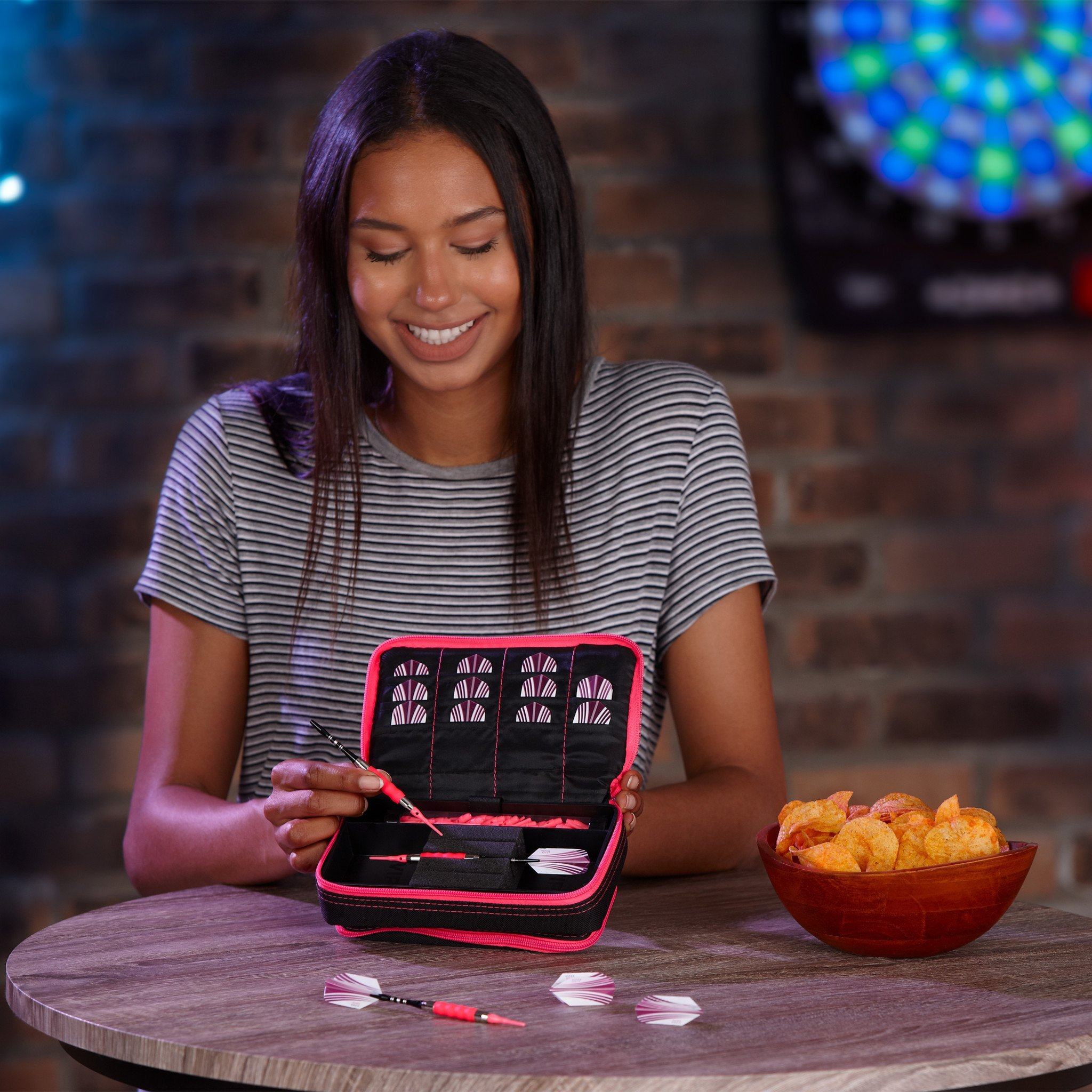 Person examining open black dart case with pink trim and white darts on table next to bowl of snacks