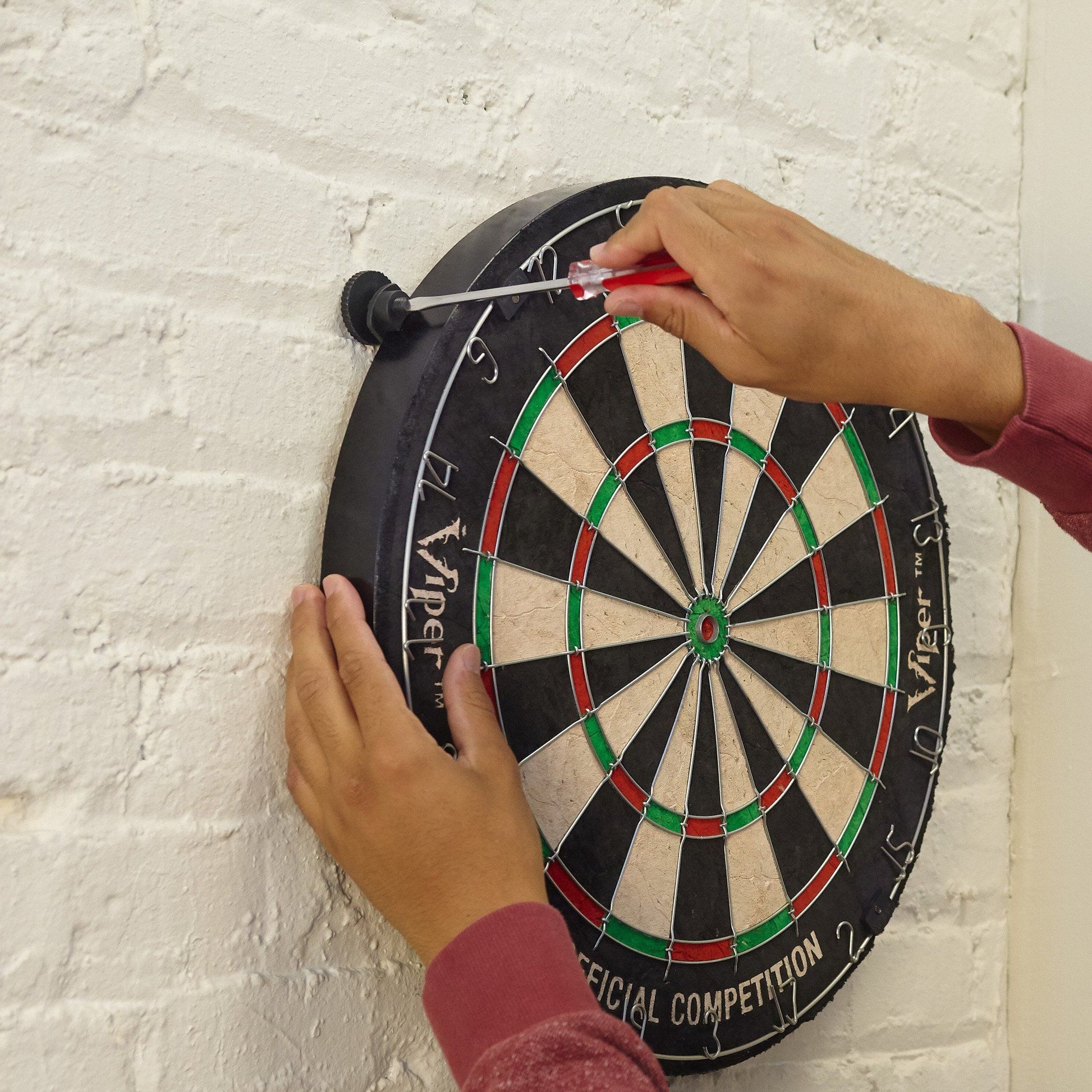 A person's hands holding a dartboard with red, green, and cream segments against a brick wall
