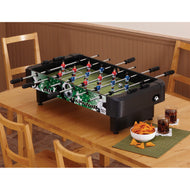 Foosball table with black frame and green playing field on wooden table with snacks and drinks beside it